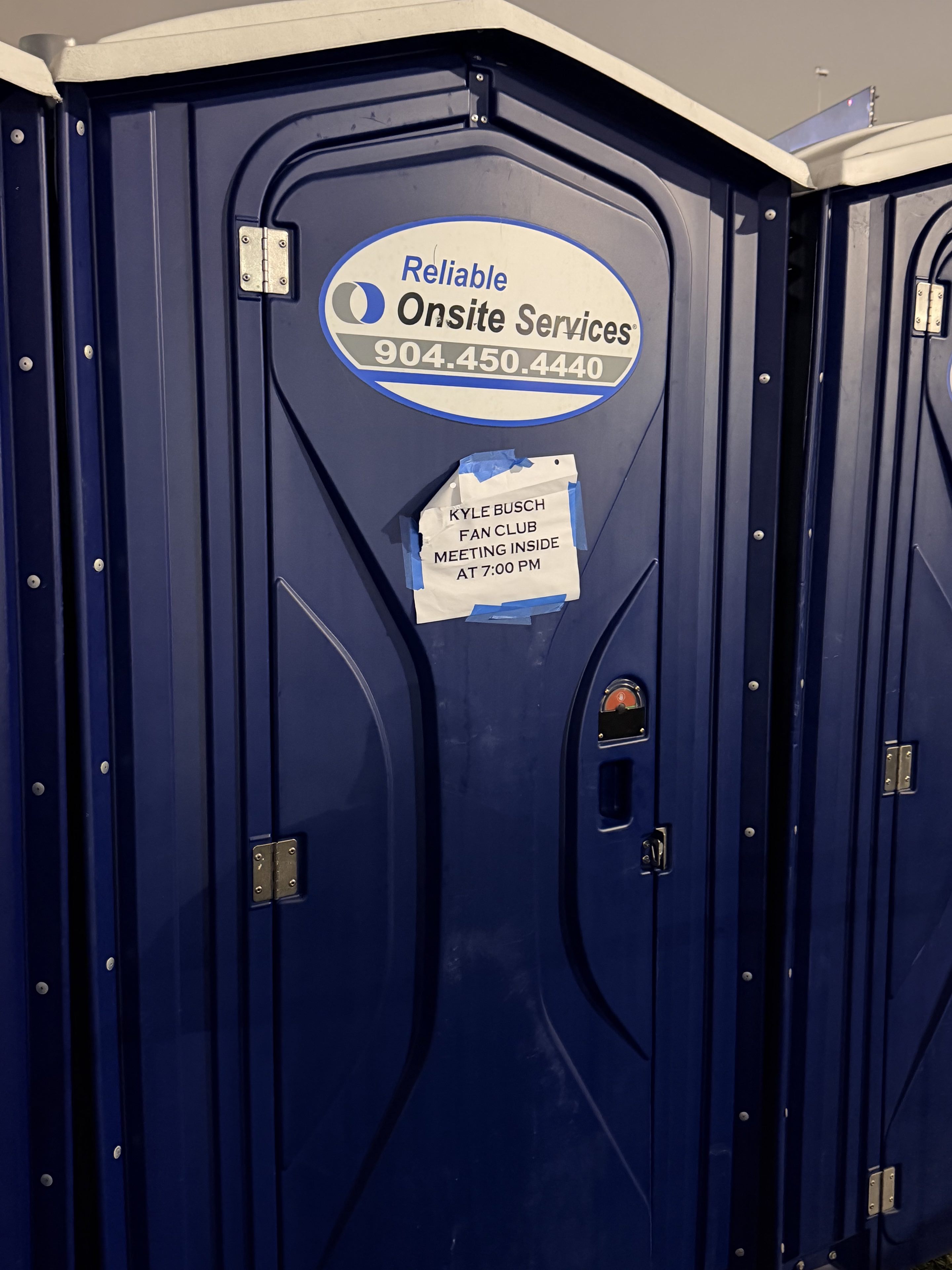 A porta potty with a sign taped on it reading “Kyle Busch fan club meeting inside at 7:00 PM”